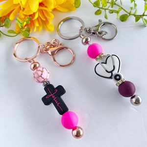 May include: Two keychains with pink and black beads. One keychain features a cross with the word "Jesus" on it. The other keychain features a white stethoscope with a heart and rhinestones. Both keychains have silver and gold metal rings.