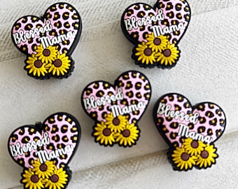 May include: Five pink and black heart-shaped shoe charms with a leopard print design. Each charm has the text "Blessed Mama" and a cluster of three yellow sunflowers.