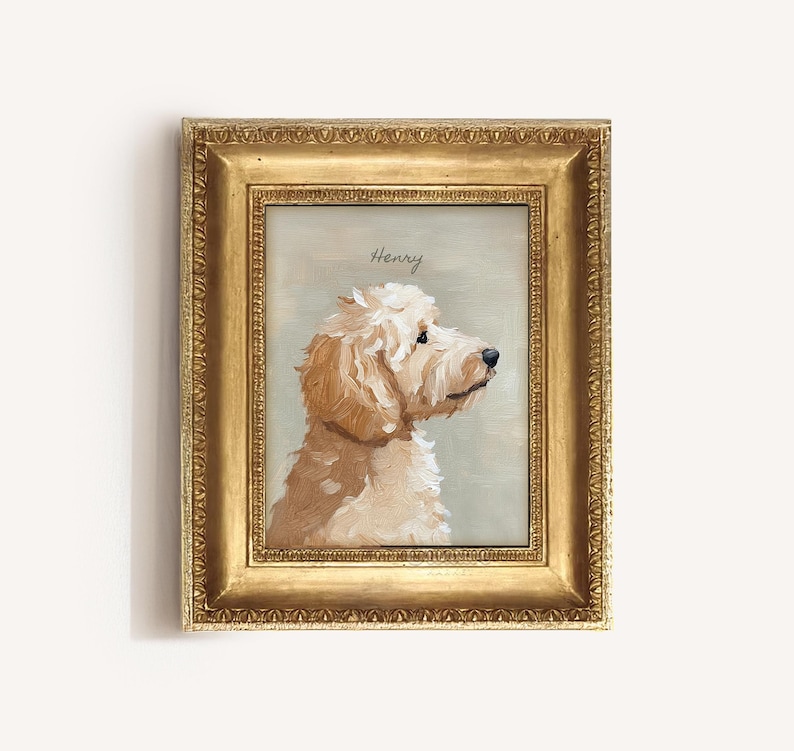 May include: A framed portrait of a dog, painted in shades of cream and tan, with the name "Henry" above the dog's head. The dog is facing right, with a fluffy coat and dark eyes. The frame is gold.