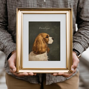May include: A framed portrait of a Cavalier King Charles Spaniel, with brown and white fur, set against a dark green background. The name "Penelope" is written in cursive at the top. The frame is gold.