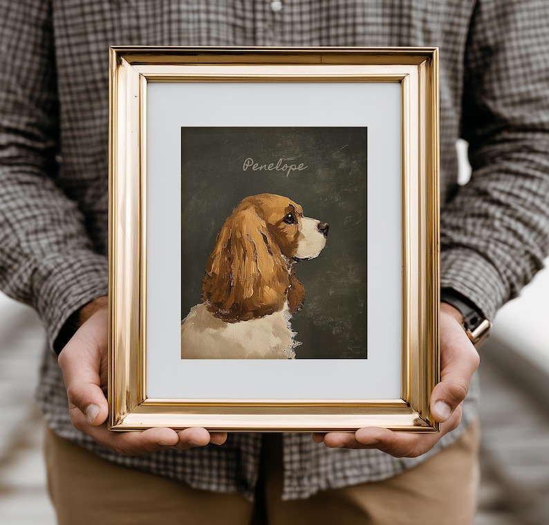 May include: A framed portrait of a Cavalier King Charles Spaniel, with brown and white fur, set against a dark green background. The name "Penelope" is written in cursive at the top. The frame is gold.