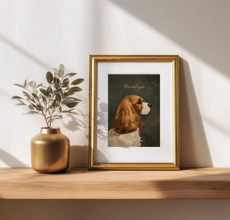 May include: A framed portrait of a Cavalier King Charles Spaniel named Penelope. The dog is depicted in profile with brown and white fur against a dark green background. The gold frame sits on a wooden shelf next to a gold vase with greenery.