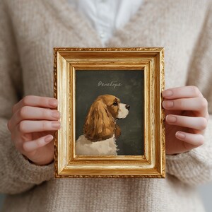 May include: A framed portrait of a dog, featuring a golden ornate frame. The artwork depicts a Cocker Spaniel with brown and white fur, set against a dark green background. The name "Penelope" is written in cursive at the top.