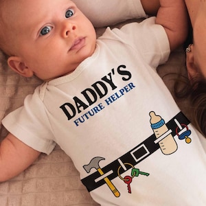 Puede incluir: Un bebé con un body blanco con el texto "DADDY'S FUTURE HELPER" en negro. El body presenta un diseño de cinturón de dibujos animados con herramientas y un biberón. Una mujer y el bebé están acostados sobre una superficie texturizada marrón claro.