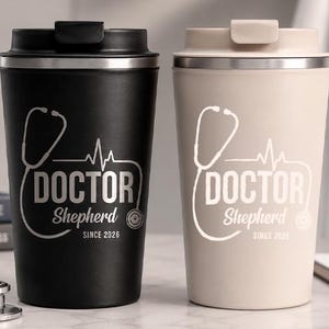 Op de afbeelding: Twee reisbekers, één zwart en één beige, met zilveren accenten. Elke beker heeft een stethoscoopafbeelding, het woord "DOCTOR", de naam "Shepherd" en "SINCE 2026". De bekers hebben zwarte en beige deksels.
