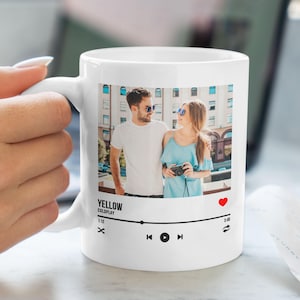 May include: White ceramic mug featuring a photograph of a couple in front of a building. The mug has a music player design with the text "YELLOW COLDPLAY" and a heart icon.