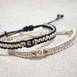Può includere: Due braccialetti dell'amicizia con cordini intrecciati neri e bianchi e perline dorate. Un braccialetto ha una perlina nera con la lettera "E" e l'altro ha una perlina bianca con la lettera "G".