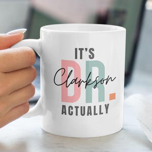 Puede incluir: Taza de cerámica blanca con un diseño rosa y verde que dice "It's Dr. Clarkson Actually".