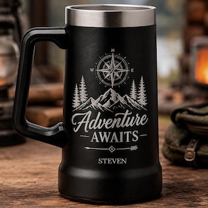 Può includere: Un boccale da birra nero opaco con bordo e manico argentati, caratterizzato da un'incisione raffigurante una bussola, una catena montuosa e alberi di pino. Il testo Adventure AWAITS e il nome STEVEN sono incisi sotto una freccia frontale.