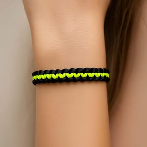 May include: A black and neon yellow woven bracelet is shown on a person's wrist. The bracelet is made of a woven material with a zig-zag pattern of neon yellow thread running through the center. The bracelet is a simple, yet stylish accessory.