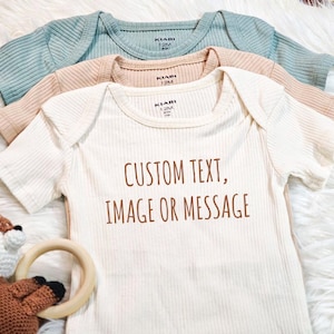 Puede incluir: Un body de bebé blanco de canalé con el texto "CUSTOM TEXT, IMAGE OR MESSAGE" impreso en marrón. El body está superpuesto a otros dos bodies, uno beige y otro azul claro.