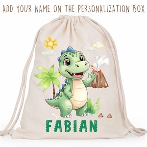 Può includere: Una borsa con coulisse beige con un'illustrazione di un simpatico dinosauro verde, un vulcano, una palma e un sole. Il nome "FABIAN" è stampato in verde. Il testo in alto recita "ADD YOUR NAME ON THE PERSONALIZATION BOX."