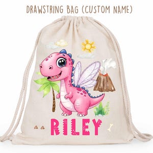 Può includere: Una borsa con coulisse beige con un'illustrazione di dinosauro rosa, ali di fata e il nome "RILEY" in lettere rosa. La borsa presenta anche una palma, un vulcano, un sole e nuvole. Il testo "DRAWSTRING BAG (CUSTOM NAME)" è in alto.