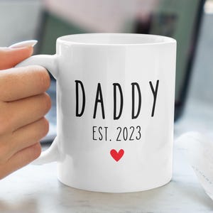 Puede incluir: Taza de cerámica blanca con texto negro que dice "Daddy Est. 2023" y un pequeño corazón rojo.