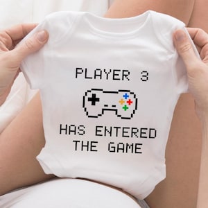 Op de afbeelding: Witte baby romper met de tekst "PLAYER 3 HAS ENTERED THE GAME" en een gepixelde afbeelding van een videogamecontroller.