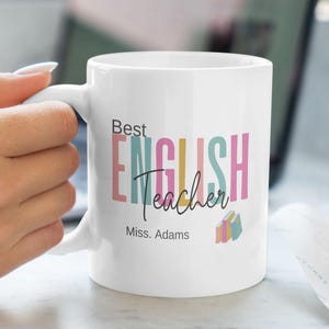 Puede incluir: Taza de cerámica blanca con un diseño colorido que dice "Best English Teacher" y "Miss. Adams" en la parte inferior. La taza tiene un esquema de color rosa, azul y verde.