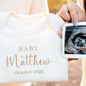 Puede incluir: Body de bebé color crema con el nombre "Matthew" y el texto "October 2025" impresos en escritura color cobre. Una imagen de ultrasonido se sostiene junto al body, revelando un bebé. El body es sostenido por una persona.