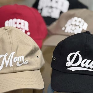 Op de afbeelding: Vier baseballpetten in verschillende kleuren en met tekst. De petten zijn beige, zwart, rood en grijs. De tekst op de petten is "Mom", "Dad", "Soccer" en "Soccer Mom".