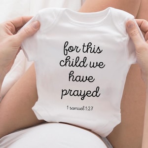 Könnte beinhalten: Weißer Baby Strampler mit schwarzem Text, der "for this child we have prayed" und "1 Samuel 1:27" lautet.