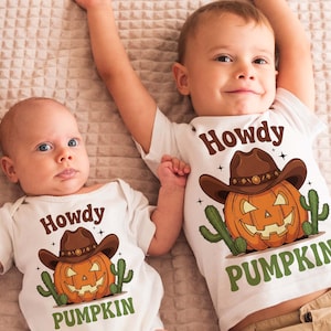Puede incluir: Dos bebés con camisetas y bodies blancos con un diseño temático de Halloween. El diseño presenta una calabaza sonriente con sombrero de vaquero, con las palabras "Howdy Pumpkin" en marrón y verde.
