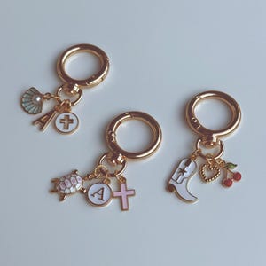 Pode incluir: Três chaveiros dourados com vários pingentes. Um chaveiro tem uma concha, uma cruz e a letra "A". Outro tem uma tartaruga, uma cruz e a letra "A". O terceiro tem uma bota, um coração e cerejas.