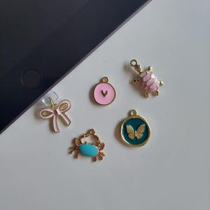 May include: Five gold charms with enamel details. The charms are a pink bow, a pink turtle, a blue crab, a green butterfly, and a pink heart with a white letter V.