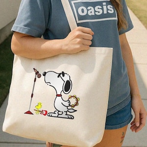 Może przedstawiać: Kremowa torba typu tote z kreskówkowym Snoopy grającym na tamburynie, z Woodstockiem i gitarą. Torba ma biały pasek. Osoba ma na sobie niebieski t-shirt z napisem "oasis".