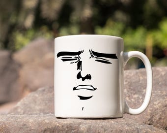 Puede incluir: Taza de cerámica blanca con un rostro en negro, con los ojos cerrados y una expresión neutra. La taza tiene un asa curvada. El fondo es borroso, con elementos naturales.