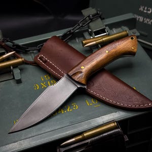 May include: A brown leather sheath with a stitched design holds a hunting knife with a wooden handle and a silver blade. The knife is resting on a green metal box with yellow markings.