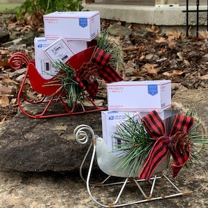 May include: Two miniature red and silver sleighs decorated with pine sprigs, red and black plaid ribbon, and pine cones. Each sleigh is filled with small white boxes labeled "Priority Mail" and "United States Postal Service".