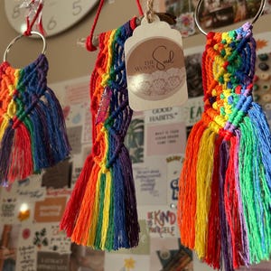 Puede incluir: Tres llaveros de macramé arcoíris con flecos. Los llaveros están hechos con hilo de colores y están unidos a anillos plateados. Los llaveros están colgados de una rama de madera.