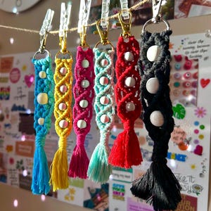 May include: A set of six colourful macrame keychains with white beads and tassels. The keychains are hanging from clips on a string.