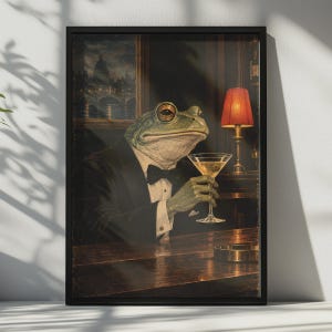 May include: A framed wall art print depicting a green frog dressed in a black tuxedo with a bow tie, sitting at a dark wooden bar. The frog holds a martini glass with an olive. A red lamp illuminates the scene, and a city view is visible behind.