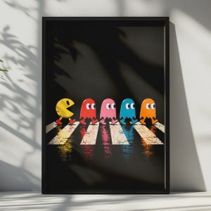 May include: A framed wall art print featuring a yellow Pac-Man character and four colourful ghosts in red, pink, blue, and orange walking in a line across a white crosswalk on a black background, with reflections visible on the ground surface.