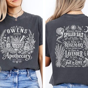 Puede incluir: Una camiseta gris carbón con estampados gráficos en blanco. El frente muestra un mortero con mano y el texto Owens Sisters Apothecary Est 1995. La parte posterior presenta ilustraciones botánicas y texto sobre sal, romero y lavanda.
