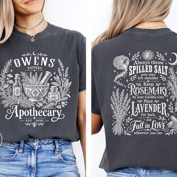 May include: A charcoal gray t-shirt featuring white graphic prints. The front displays a mortar and pestle with the text Owens Sisters Apothecary Est 1995. The back features botanical illustrations and text about salt, rosemary, lavender, and love.
