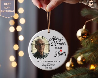 May include: A white ceramic memorial ornament with a photo of a man in glasses. Text reads "Always & Forever in our Hearts" and "In Loving Memory of Joseph Stewart 1938-2023". The ornament is held by a gold string.