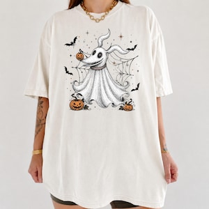 May include: An off-white short-sleeve t-shirt featuring a black and white graphic illustration of a ghost dog with long ears. The design includes orange jack-o'-lantern pumpkins, black bats, and a spider web background with small star accents.