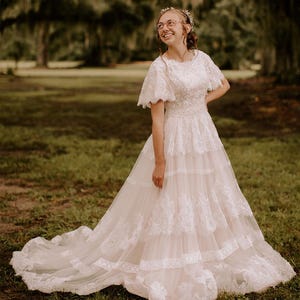 May include: A woman wearing a white lace wedding dress with a tiered skirt and a short sleeve design.