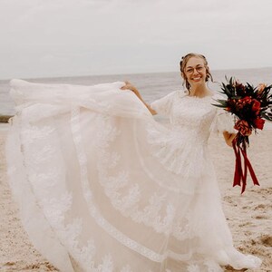 May include: A white lace wedding dress with a fitted bodice and a full skirt. The dress has a scoop neckline and short sleeves. The bride is holding a bouquet of red and green flowers.