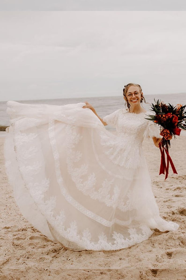 May include: A white lace wedding dress with a fitted bodice and a full skirt. The dress has a scoop neckline and short sleeves. The bride is holding a bouquet of red and green flowers.