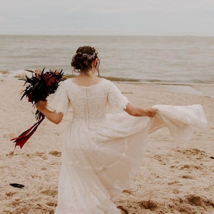 May include: A bride in a white lace wedding dress with a bouquet of red and green flowers walks on a sandy beach.