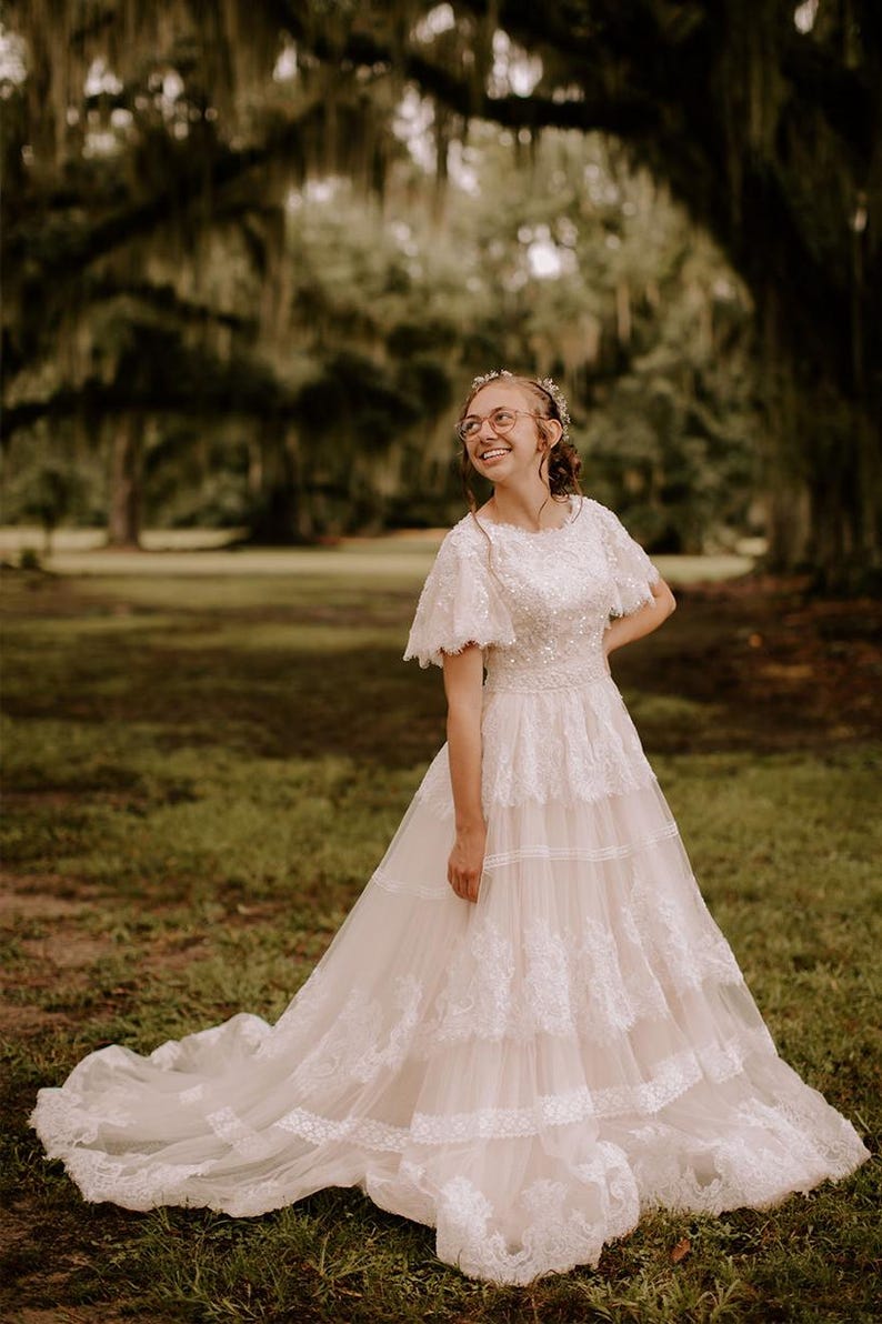 May include: A woman wearing a white lace wedding dress with a tiered skirt and a short sleeve design.