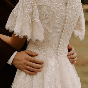 May include: A white lace wedding dress with a beaded bodice and a tulle skirt. The dress has a scoop neckline and short sleeves. The dress is buttoned up the back.