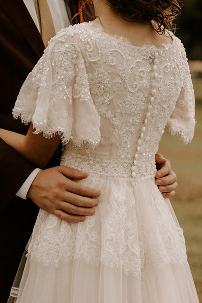 May include: A white lace wedding dress with a beaded bodice and a tulle skirt. The dress has a scoop neckline and short sleeves. The dress is buttoned up the back.