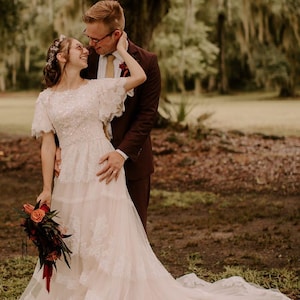 May include: A bride and groom standing in a grassy field. The bride is wearing a white lace wedding dress with a long train. The groom is wearing a dark brown suit. The bride is holding a bouquet of red and orange flowers. The couple is looking at each other and smiling.
