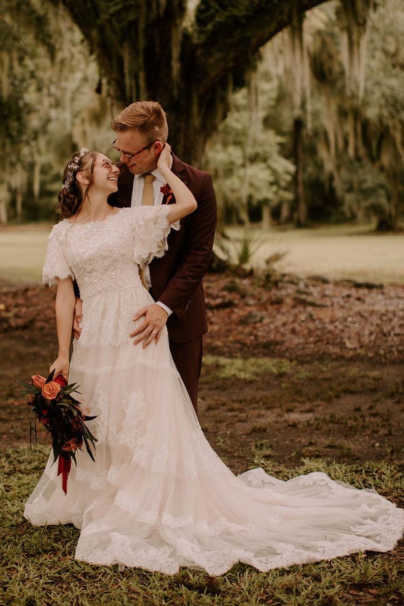 May include: A bride and groom standing in a grassy field. The bride is wearing a white lace wedding dress with a long train. The groom is wearing a dark brown suit. The bride is holding a bouquet of red and orange flowers. The couple is looking at each other and smiling.