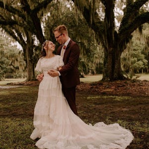 May include: A couple poses for a wedding photo in front of a large tree with Spanish moss. The bride is wearing a white lace dress with a long train. The groom is wearing a burgundy suit and a white shirt. The couple is smiling and looking at each other.