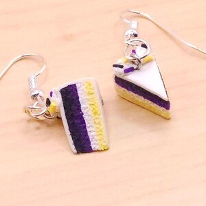 May include: Silver-tone earrings with cake slice charms. Each charm has a white top, purple and yellow layers, and a small purple decoration. The earrings are designed with a hook style for pierced ears. The cake slices are miniature.