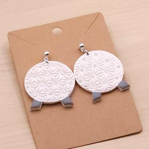 May include: A pair of white, round earrings with a geometric pattern and small, clear embellishments. The earrings have silver-colored posts and gray, rectangular accents at the bottom. They are displayed on a brown card.